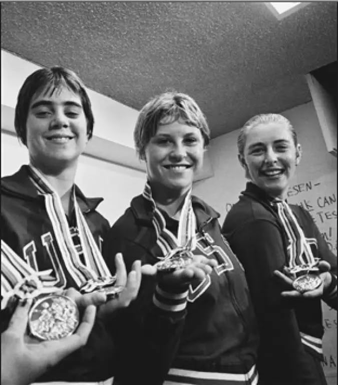 Black-and-white photo of three smiling U.S. swimmers in team jackets holding up their Olympic medals
